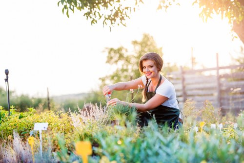 A woman with dark hair tied back, wearing a white T-shirt and dark apron, is tending to a garden in daylight, surrounded by a variety of flowering plants, shrubs, and small trees. She is smiling while using a garden tool to trim or prune the plants. The garden features a well-maintained lawn, flower beds with colorful blooms, and a wooden fence in the background. The environment appears sunny with soft natural light, indicating clear weather. The scene is set in an outdoor space that could be part of a residential garden in Crouch End, London, highlighting typical landscaped and planted areas that require regular gardening and outdoor maintenance services provided by professionals such as Landscapers Crouch End.