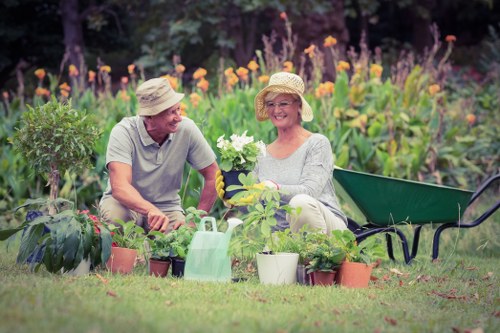 A gardener and a woman, both wearing wide-brimmed straw hats, are seated in a lush, well-maintained garden with vibrant flowering plants and shrubs in the background. The gardener, dressed in a light grey T-shirt, is smiling while tending to potted plants, including small flowering and leafy species arranged on the grass in front of them. The woman is holding a white flowering plant, enjoying the outdoor space which features a neatly trimmed lawn, a black garden bench nearby, and a watering can among the pots. The scene is set in bright daylight with clear, natural shadows, in an outdoor garden area that emphasizes the care and upkeep of the landscaped garden, which could be part of a residential back garden in Crouch End or the surrounding area. This image reflects gardening activities and outdoor plant management typical of professional garden maintenance services in London, highlighting their expertise in garden cultivation and landscaping.