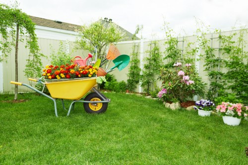 A well-maintained backyard garden with a vibrant green grass lawn, bordered by a white composite fence covered in climbing plants and greenery. In the center of the scene, a yellow wheelbarrow filled with colorful flowers and gardening tools such as a pink watering can and orange trowel. To the right, there are neatly arranged flower beds containing blooming pink and purple flowers, with some potted plants placed along the edge. In the background, leafy trees and shrubs frame the outdoor space, with the roof of a neighbouring house visible above the fence. The environment appears bright and sunny, suggesting a clear day suitable for gardening activities. This outdoor area reflects a typical landscaped garden that could benefit from professional gardening and landscaping services offered by Landscapers Crouch End, particularly for garden maintenance, flower planting, and lawn care, fitting within the local Crouch End community in North London.