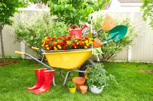 In a lush, well-maintained front garden in Crouch End, London, a bright yellow wheelbarrow filled with vibrant red and yellow flowering plants sits on a neatly trimmed lawn. Surrounding the wheelbarrow are various gardening tools and supplies, including a red watering can, a pair of red rubber boots, and several empty terracotta pots. To the right, there's a white pot with lush green foliage, and a small yellow container. In the background, a white fence encloses the garden space, with mature trees and dense shrubbery providing a natural, green backdrop. The scene suggests active outdoor gardening in a residential outdoor area during a sunny day, highlighting garden maintenance and landscaping activities typical for local garden care in Crouch End. This setting emphasizes the importance of lawn care, plant cultivation, and outdoor aesthetic improvement as part of professional gardening services offered by Landscapers Crouch End.