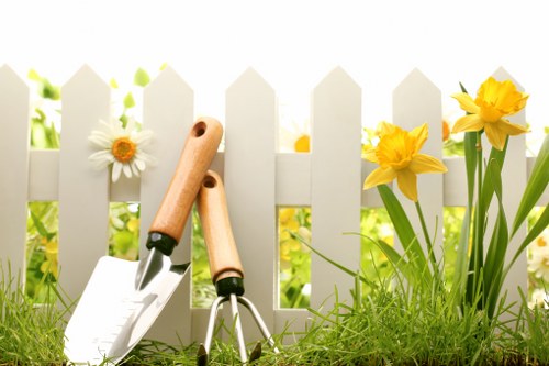 A close-up view of a tidy garden border in a residential outdoor space, featuring bright yellow daffodils and white daisies blooming alongside a white picket fence. The foreground shows well-maintained, lush green grass with a pair of small gardening tools, including a trowel and a hand rake, laid on the ground. Behind the fence, soft natural light illuminates the scene, suggesting a clear, sunny day. The area appears to be part of a front or back garden in Crouch End, with the tools indicating recent gardening activity. The combination of flowering plants, neat grass, and a white picket fence presents a welcoming, cared-for outdoor environment suitable for gardening services offered by companies like Landscapers Crouch End, focusing on planting, lawn care, and garden maintenance. The scene highlights a typical small garden setup within a residential area, emphasizing natural tones, plant variety, and tidy surfaces that form the basis for professional landscaping improvements.