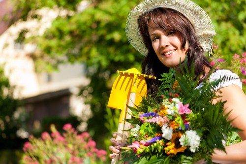 A woman in a sunlit garden holding a colourful bouquet of flowers and a yellow garden tool, with lush green foliage, blooming plants, and a paved patio visible in the background. The garden features a neatly maintained lawn with dense grass, bordered by a variety of flowering plants and shrubs, some with pink and white blossoms. She is wearing a light-colored sun hat and a white top, suggesting a pleasant outdoor setting typical of residential gardens in Crouch End. The scene appears well-maintained, reflecting professional gardening services such as lawn care, planting, and outdoor landscape maintenance by Landscapers Crouch End. Natural daylight highlights the vibrancy of the flowers and greenery, indicating good weather conditions, and the overall environment exudes a tidy, aesthetically pleasing outdoor space designed for relaxation and outdoor enjoyment.