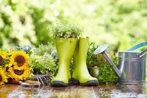 The image shows a garden scene with a wooden outdoor surface in the foreground, featuring a pair of bright green rubber gardening boots filled with white and green flowering plants, positioned centrally. To the left, there is a bouquet of vibrant yellow sunflowers mixed with purple and white flowers, while on the right, a metal watering can with a green hose attachment is placed. The background reveals a lush, out-of-focus garden with dense green foliage, suggesting a well-maintained outdoor space typical of a suburban garden in Crouch End or the surrounding London area. The scene is illuminated by natural daylight, indicating fair weather, and evokes a neat, inviting environment suitable for gardening activities, aligning with the services provided by Landscapers Crouch End in lawn, planting, and outdoor maintenance for residential gardens.