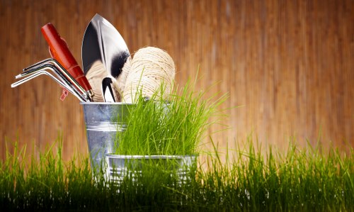 A close-up view of a small garden with a vibrant green lawn in the foreground, featuring neatly trimmed grass with a mix of short and slightly taller blades, indicative of regular maintenance. Behind the grass, there is a rustic wooden garden fence with a warm brown tone, providing a natural boundary for the outdoor space. The background includes a few potted plants and garden decorations, suggesting a well-tended residential garden in Crouch End. The overall setting exudes a calm and tidy outdoor environment suitable for gardening activities, landscaped to promote sustainability and eco-friendly outdoor care, aligning with services at landscaperscrouchend.co.uk. The lighting suggests a bright, overcast day, casting soft, even light on the garden surfaces, perfect for appreciating the natural textures of the lawn, wooden elements, and greenery.
