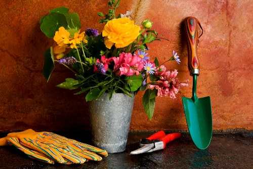 A close-up of a garden workspace setup, featuring a large pot filled with colorful flowers including yellow, pink, and purple blooms, sitting on a dark stone surface against a reddish-brown wall. Next to the pot are a pair of gardening gloves with yellow and green stripes, a small red hand rake, a pair of pruning shears, and a green-handled trowel with a wooden grip. The background suggests an outdoor or patio garden area, ready for planting or maintenance work, with natural lighting highlighting the vibrant flower colors and garden tools. This scene reflects outdoor gardening preparations, landscaping tasks, and plant care practices typical of a well-maintained garden space in Crouch End, highlighting the importance of proper tools and equipment for garden enhancement and landscaping projects.