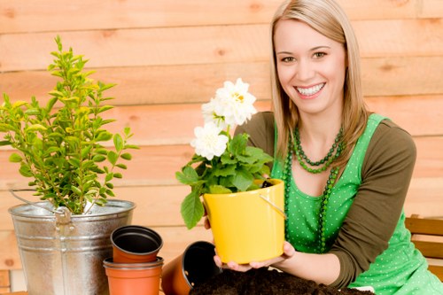 The image depicts a woman with shoulder-length blonde hair smiling as she holds a vibrant yellow flowerpot with white flowering plants, situated in a garden or outdoor patio setting. In front of her are various gardening tools and pots, including a large metal bucket with a lush, green shrub or small tree, and several empty pots placed on the surface. The background features a wooden fence or wall, suggesting a private garden space typical of a residential outdoor area in Crouch End. The scene is well-lit with natural daylight, indicating good weather conditions, and the environment appears neat and well-maintained, reflecting gardening activities. The woman’s attire, casual yet cheerful, aligns with gardening and outdoor maintenance themes, supporting landscaping and garden revitalisation services offered by Landscapers Crouch End in the local area.