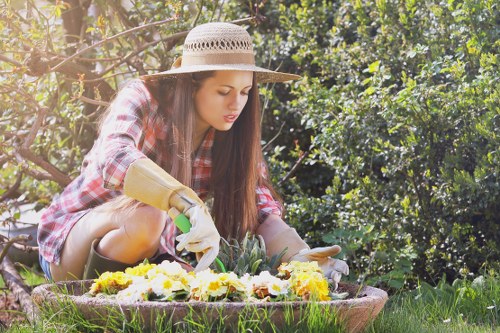 A young woman wearing a wide-brimmed straw hat, a plaid shirt, and gardening gloves is kneeling on a lawn in a garden, carefully tending to a circular flower bed filled with a variety of blooming yellow, white, and purple flowers. The garden features a neatly maintained grassy area with dense green hedges and trees in the background, providing natural shade and privacy. The scene is lit by natural daylight, illustrating a peaceful outdoor space ideal for garden renovation and landscaping projects near Crouch End, London. The woman’s focused activity highlights outdoor gardening and maintenance, relevant to services offered by Landscapers Crouch End in the local area, with clear visibility of garden elements like soil, flower beds, and lush greenery.