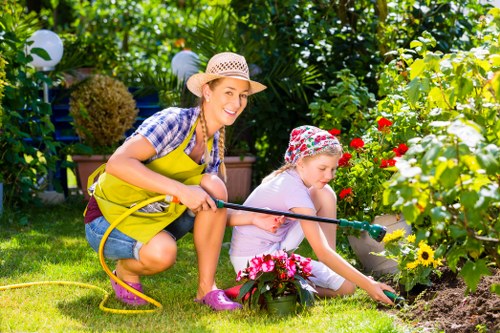A woman and a young girl are engaged in gardening activities in a well-maintained backyard garden. The woman, wearing a straw hat, a blue checkered shirt, and a bright yellow apron, is smiling as she crouches beside the girl and holds a garden hose, which she is using to water the plants. The girl, dressed in a pink shirt and floral hat, is focused on planting or tending to a flower bed bordered with pink and red flowers. The garden features lush green grass, a variety of flowering plants, and a backdrop of dense foliage including bushes and small trees. The sunlight filters through the leaves, creating bright and shadowed areas, indicating a clear day. Decorative garden lanterns hang from the surrounding plants, adding to the inviting outdoor environment. This scene depicts typical gardening and outdoor maintenance activities suitable for a landscaped yard in the Crouch End area, emphasizing natural tones, plant diversity, and garden care. Landscapers Crouch End supports maintaining such outdoor spaces for local homes.