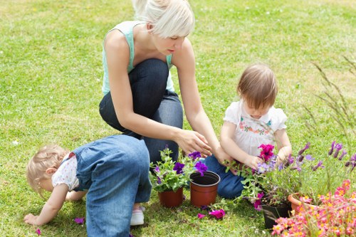 A woman with short blonde hair, wearing a light blue sleeveless top and dark jeans, is kneeling on a well-maintained lawn in a residential garden, tending to potted plants and garden flowers. Two young children, a girl with brown hair in a white top with floral patterns and blue jeans, and another child in denim overalls, are engaged in outdoor gardening activities, with the girl holding a small plant and the other child exploring the grass. The garden features a lush, green grassy area with a border of flowering plants in shades of purple, pink, and orange, along with pots placed on the soil for planting. In the background, there are neatly trimmed hedges and a paved pathway, suggesting an organized and cared-for outdoor space typical of a suburban garden in Crouch End, London. The scene is lit by natural daylight, indicating a mild, sunny day suitable for gardening activities, reflecting professional outdoor maintenance and gardening services offered by Landscapers Crouch End.