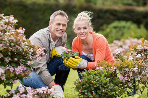 A man and a woman are working together in a lush garden, surrounded by flowering shrubs with pink and white blooms. The man, dressed in a light-colored shirt, and the woman, in an orange jacket, are both smiling at the camera while wearing yellow gardening gloves. They are crouched near a variety of well-maintained bushes and plants, indicating active gardening or pruning. Behind them, the garden features a neatly trimmed lawn area with dense greenery and trees in the background, suggesting an outdoor residential space typical of Crouch End or nearby London suburbs within N8 postcode. The scene is set on a bright, partly cloudy day, with natural light highlighting the vibrant colors of the plants and the gardeners' clothing. This image reflects the professional gardening services offered by Landscapers Crouch End, focusing on lawn maintenance, shrub trimming, and garden enhancement in the local area, emphasizing the importance of well-kept outdoor spaces in urban gardens.